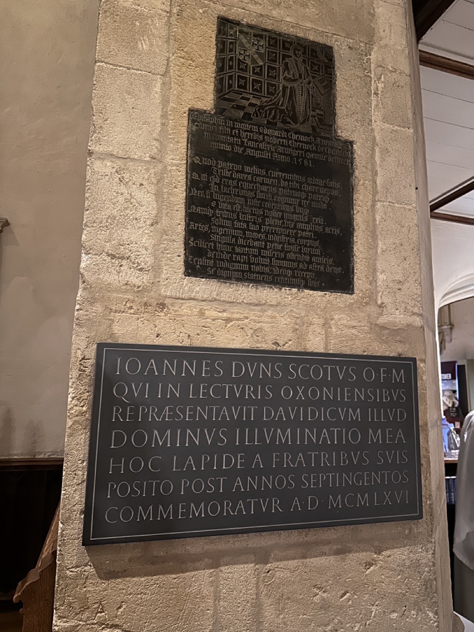 Two separate memorials on a stone pier: Edward Chernock’s figurative brass above a Latin plaque commemorating John Duns Scotus.