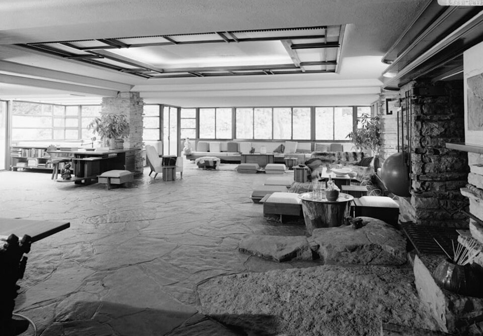 Fallingwater living room seen from the kitchen, with the projecting hearth rock at right, desk at left, perimeter seating and a ceiling light screen overhead.
